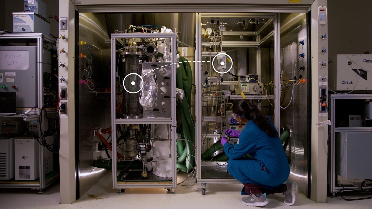 A researcher kneels beside a walk-in fume hood, where scientific equipment stands.