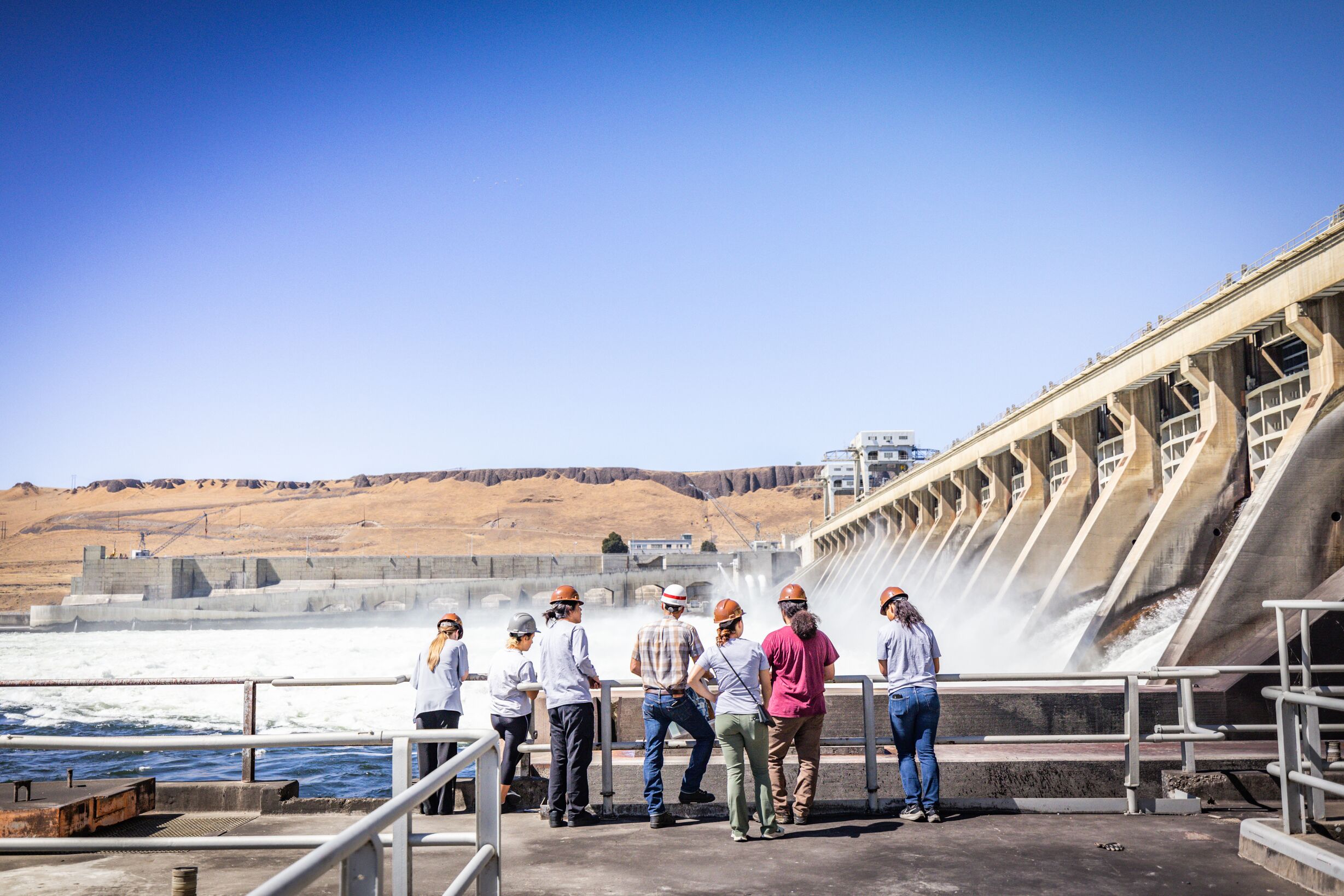 Photo of people in safety helmets, outside, observing the McNary Dam.