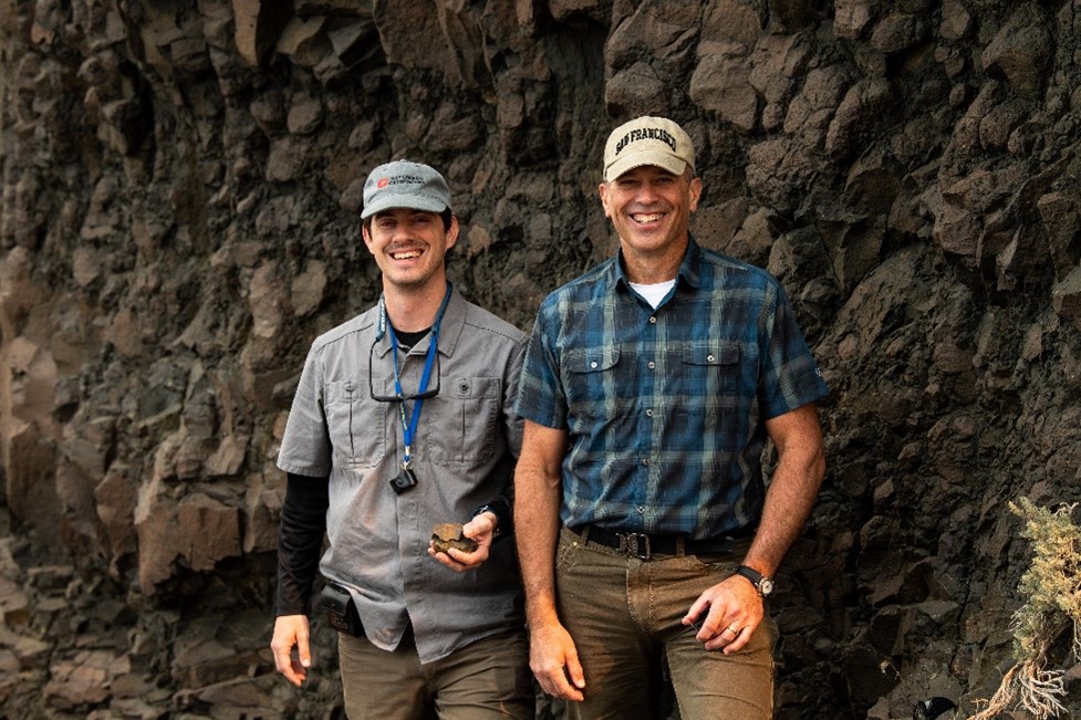 Two men wearing hats and outdoor clothing in front of a rocky cliff