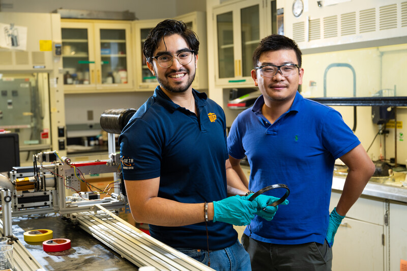 Portrait photo of Cesar Moriel and his mentor Yao Qiao inside a lab room, wearing safety gloves