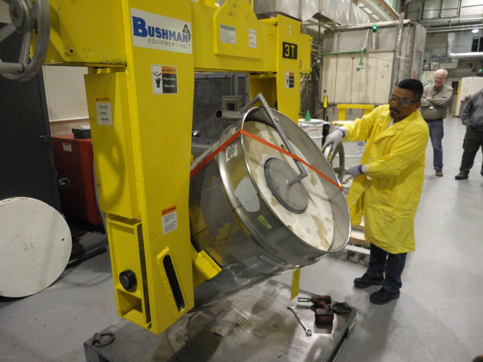 A man in a yellow lab coat uses equipiment to rotate a large metal drum
