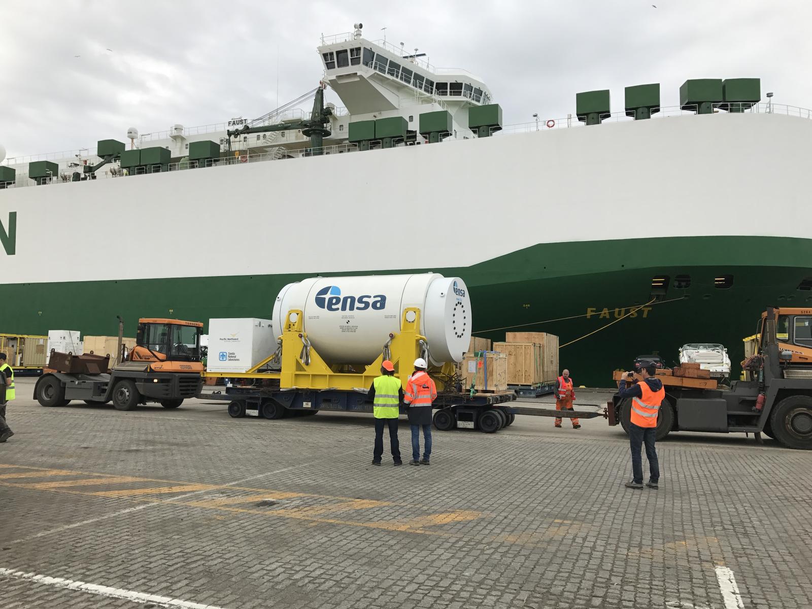 A large white cylinder sits on a yellow platform, waiting to be lifted by crane onto a ship.