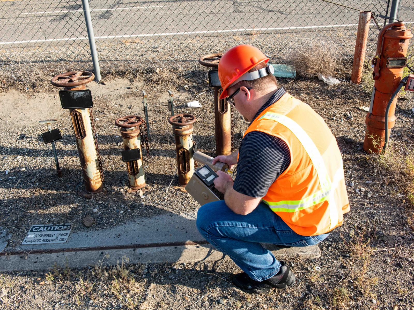 Man wearing safety vest and hard hat holds a geiger counter by some rusty pipes to see if they are radioactive