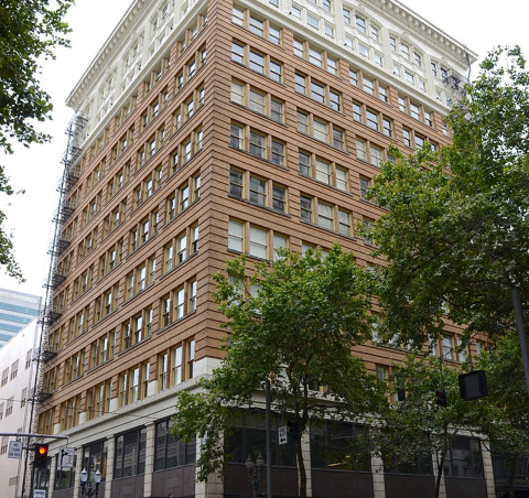 The PNNL Portland Office, a square red brick building with evenly spaced windows on each floor, pictured from street level with green trees in the foreground