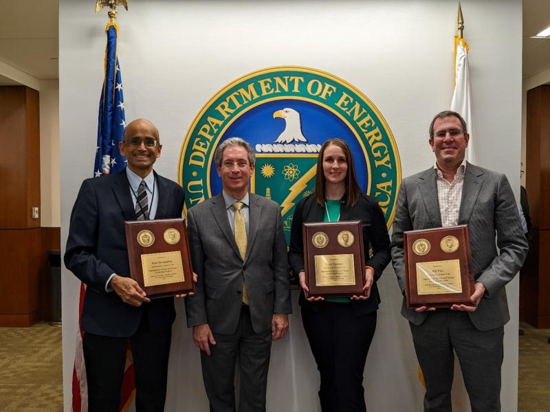 Four people standing in front of a U.S. Department of Energy logo. Three are holding plaques.