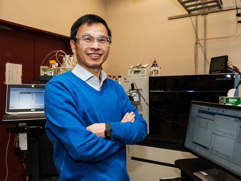 PNNL researcher Wei-Jun Qian stands near laboratory computers, smiling.