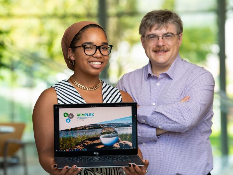 High school intern Jordan Perkins holds a laptop showing the RemPlex website, standing next to mentor researcher Chris Johnson.