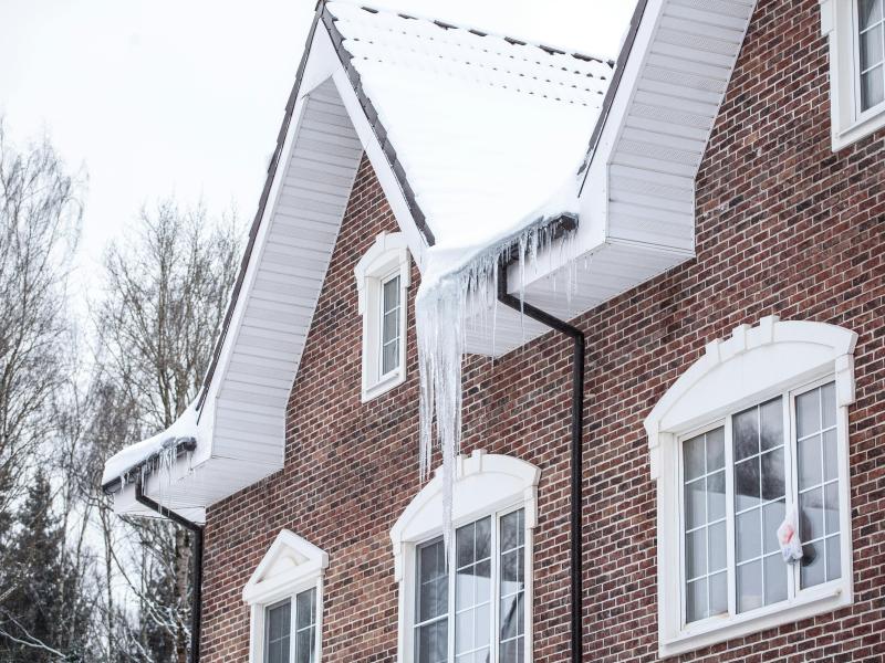 Photograph shows a brick house with a sloping roof in winter. There are icicles hanging off the edge of the roof.