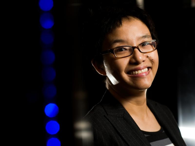 Smiling Asian woman with short hair and glasses and a dark background with blue lights